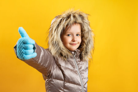 The child shows a thumb up. Happy cute little girl in a down jacket on a yellow background. Winter...