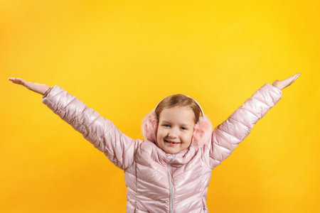 The child in a jacket and warm headphones raised her hands up. Little girl on a yellow background. The concept of happiness, winter, autumn.の写真素材