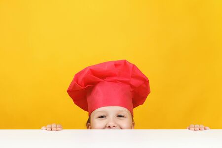 Funny little girl chef on a yellow background. The child is hiding and looking out from under the table.の写真素材