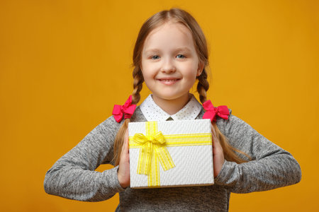 Cute little girl holding a box with a gift in her hands. Baby with pigtails on a yellow background.