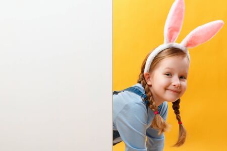 Easter concept. Cute little baby girl with bunny ears peeks out from behind a blank banner. Child hunts for eggs.の写真素材