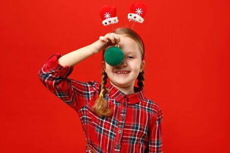 Funny happy little girl with christmas ball decoration. A child in a red dress and mittens headband on a colored background.の写真素材
