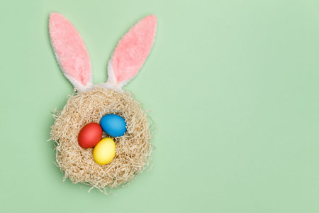 Nest with three easter eggs and bunny ears on a mint background. Top view. Copy space.