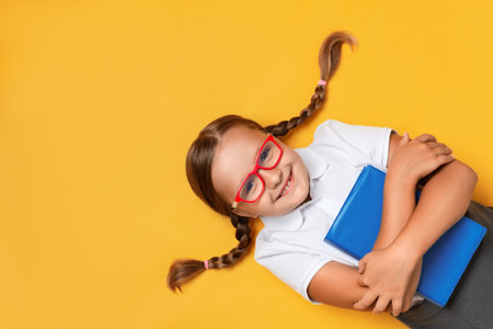 Cute little girl schoolgirl in glasses with a book on a yellow background. Top view.