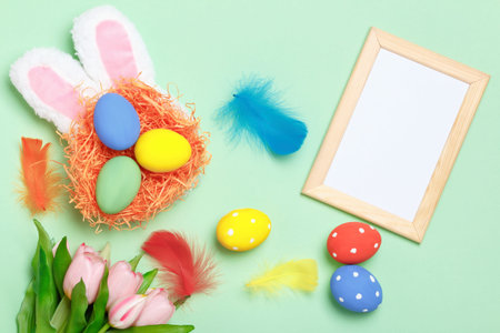 Easter eggs in a nest, bunny ears, flowers and a frame with copy space on a mint green background.