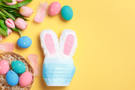 Easter concept. Rabbit in a protective medical mask and a basket with colored eggs on a yellow...