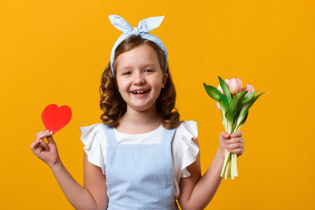 Cute happy little girl on a yellow background. The child is holding a bouquet of tulips and a paper heart.の写真素材