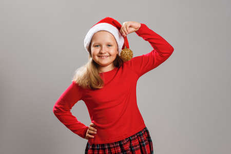 Happy child is holding a Christmas ball toy. Close-up of a little girl in a santa hat on a gray isolated background.の写真素材
