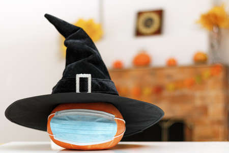 Halloween pumpkin in a witch hat in a protective mask on the table against the background of a home fireplace with autumn decorations.の写真素材