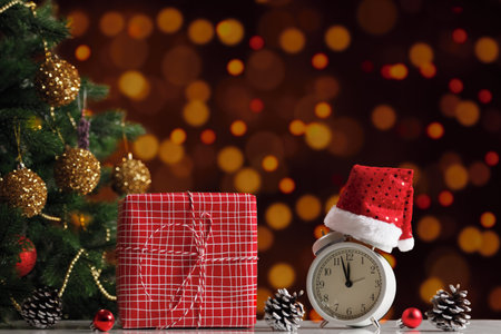 New Year's composition. Gift box and alarm clock in a Santa hat next to a decorated Christmas tree on a blurred background of lights.の写真素材