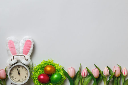 Easter concept. Alarm clock in the ears of a rabbit pearl eggs in a nest of flowers tulips on a gray...