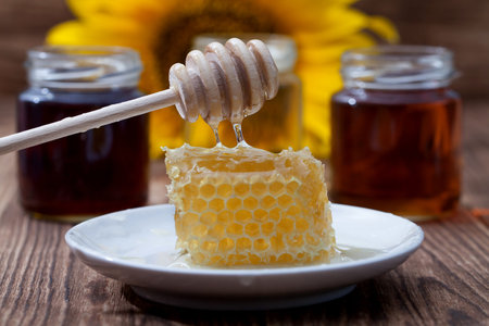 drops of honey from a spoon, honeycomb and several types of honey cans on a wooden background, focus...