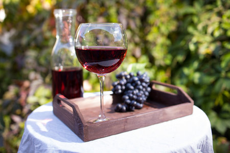 A glass of red wine prepared for tasting stands on a table with a white tablecloth outdoors in sunlightの写真素材