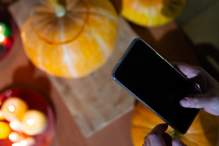 A person stands holding a cell phone in front of a table that is beautifully decorated with pumpkins for halloween, and flickering candles all aroundの写真素材