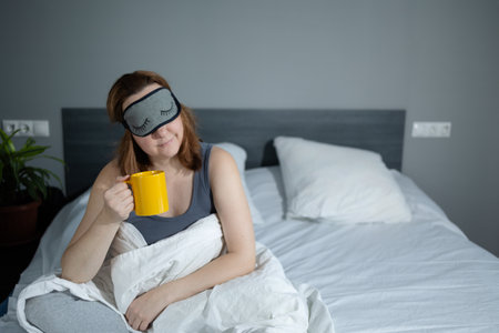 A relaxed woman in a bedroom, sipping coffee and wearing a sleep mask, embracing morning tranquility.の写真素材