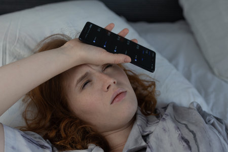 A distressed young woman lies in bed, holding a smartphone to her forehead, showing signs of fatigueの写真素材