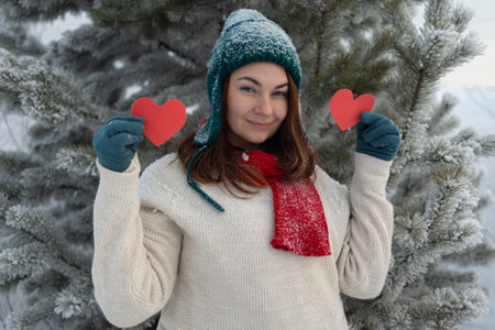 A joyful woman in a warm winter outfit holds red hearts on a snowy background, symbolizing love, unity and Valentines Day.の写真素材