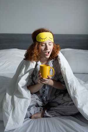 A woman happily enjoys a blissful and cozy morning while sipping coffee, wearing a soft sleep mask in bedの写真素材