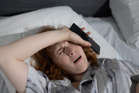 A stressed woman holds her phone tightly while lying in bed, underscoring the many challenges of modern lifeの写真素材