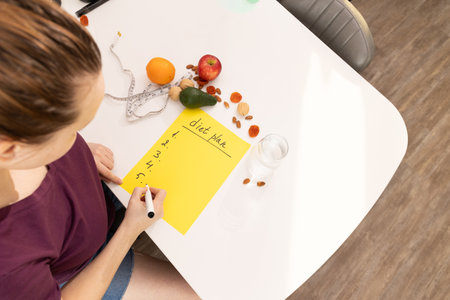 A person is carefully listing items for meal prep, surrounded by an array of fresh, colorful fruits and vegetablesの写真素材