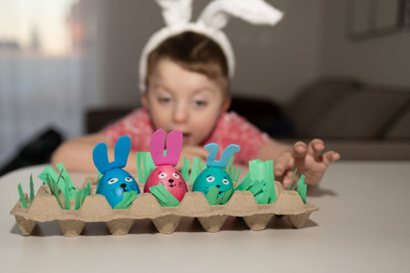 A joyful child wearing bunny ears is eagerly reaching out for colorful and vibrant Easter eggs on a tableの写真素材