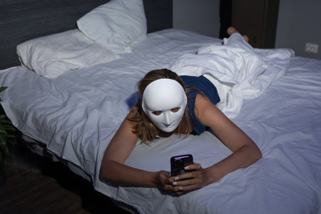In a dimly lit bedroom, a person wearing a white mask lies on their stomach on a bed while intently using a smartphone. The focus is on themes of digital privacy and data protection.の写真素材