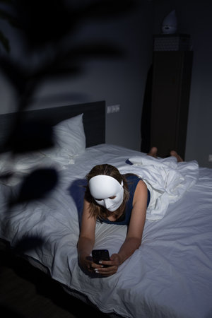 A person wearing a white mask lies on a bed, focused on their smartphone in a dimly lit bedroom. The scene highlights the importance of digital privacy and protection against data fraud.の写真素材