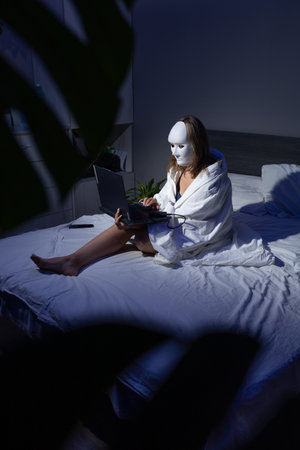 In a dimly lit room, a person draped in a blanket sits on a bed, using a laptop. Their face is obscured by a white mask, highlighting concerns over data security and fraud.の写真素材