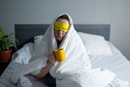 A person wrapped in a blanket, wearing a sleep mask and holding a coffee mug, enjoying a cozy morning.の写真素材