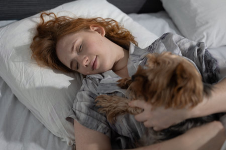 A young woman with red hair rests comfortably on a bed, holding her small dog close. The soft lighting and cozy pillows create a serene atmosphere, ideal for a restful night.の写真素材