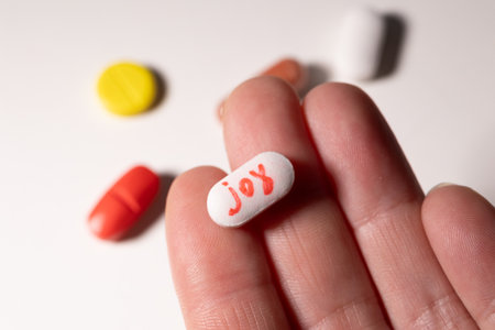 A hand holds a white pill inscribed with the word joy, surrounded by colorful pills. This representation emphasizes the complex relationship between pleasure, mental health, and addiction.の写真素材