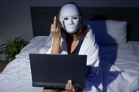 A person wearing a mask sits on a bed, draped in a blanket, while holding a laptop.の写真素材