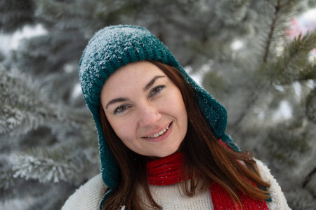A cheerful woman wearing a colorful knitted hat and a cozy scarf is smiling in a picturesque snowy...