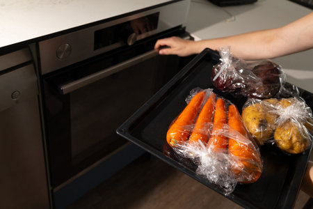 Fresh vegetables are being prepared for baking in an oven. A person holds a tray with carrots,...