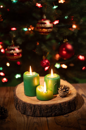 On a wooden surface, three green candles burn brightly, casting a warm glow against the backdrop of a beautifully decorated Christmas tree. Colorful lights twinkle among ornaments.の写真素材