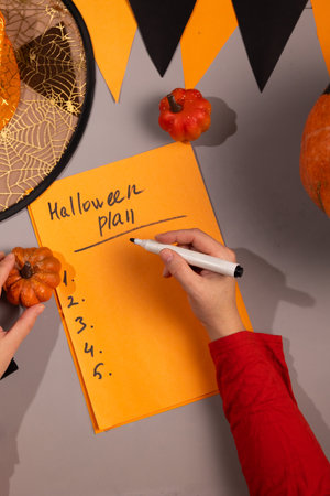 Person writes a Halloween plan on an orange paper with a marker. Pumpkins, a witch hat and Halloween banner decorate the gray surface in preparation for a party.の写真素材