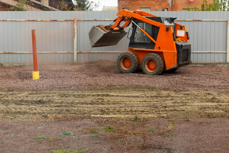 A construction vehicle is at work leveling gravel in a residential area. The action takes place during daylight, showing efforts to prepare the land for future development.の写真素材