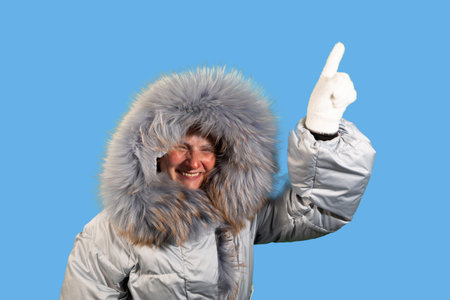 A smiling woman wearing a winter coat with a large fur hood is pointing upwards with her gloved...