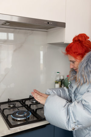 In a kitchen, a woman with red hair and a heavy winter coat warms her hands above a lit gas stove. She is trying to save money on heating and conserve energy in cold weather.の写真素材