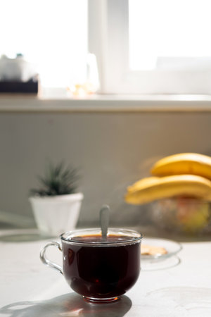 A clear glass cup filled with dark coffee rests on a kitchen counter. A spoon is in the cup. A plant, bananas, and a window are visible in the background.の写真素材