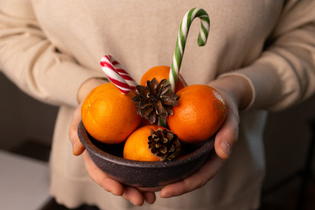 A person holds a bowl filled with oranges, candy canes, and pine cones. It suggests preparations for...