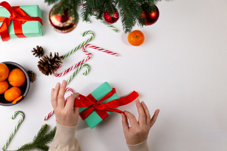 Person wrapping a gift with a bright red ribbon. There are ornaments, tangerines, and candy canes around. This is preparation for a Merry Christmas or New Year holiday.の写真素材