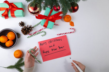 Hands writing a cheerful holiday postcard on a table decorated with Christmas gifts, oranges, and candy canes. The scene captures the spirit of Christmas and New Year preparations.の写真素材