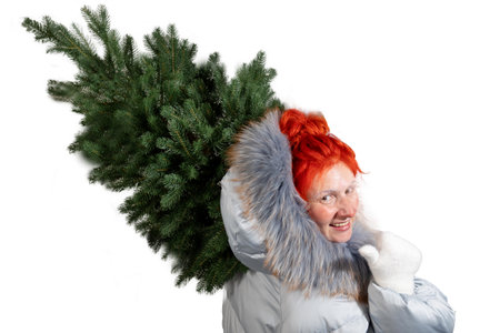 A smiling woman in winter clothes carries a green Christmas tree over her shoulder. She appears frozen with snow on her face, wearing a warm coat and gloves.の写真素材