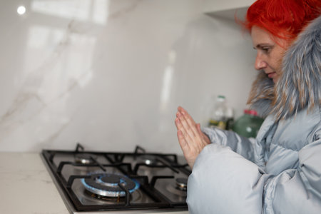 In Winter, a woman with red hair, wearing a warm puffer jacket, warms her hands over a gas stove in her kitchen. She is saving on heating costs in cold weather.の写真素材