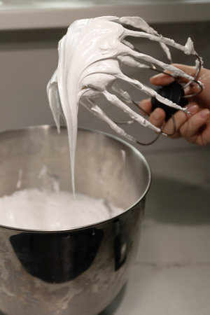 A pastry chef skillfully prepares whipped cream mixed with meringue in a shiny metal bowl, showcasing the smooth texture and glossy finish at a well-equipped cooking space.の写真素材