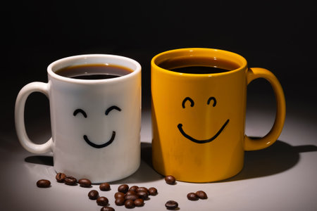 Two coffee mugs, one white and one yellow, have smiley faces drawn on them. Roasted coffee beans are scattered on the surface, suggesting a good morning start.の写真素材