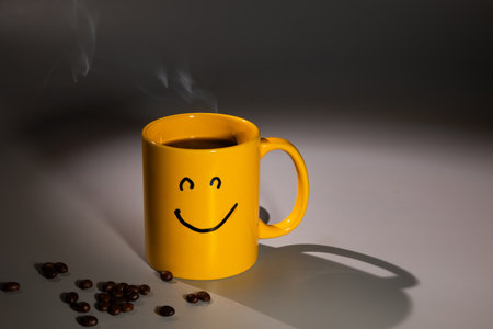 A yellow coffee mug featuring a smiley face sits on a surface, near a scattering of coffee beans. Steam rises from the mug, suggesting a warm and inviting beverage.の写真素材
