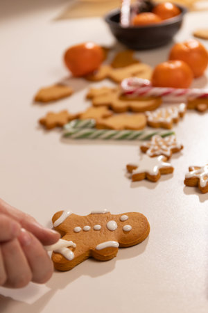 A person decorates a fresh gingerbread cookie with white icing while surrounded by other festive...