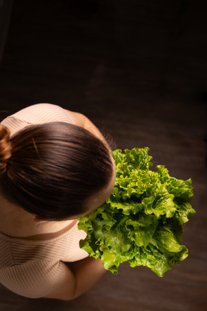 Top down view shows a woman holding a bunch of fresh green lettuce. She wears a light colored top and has her hair in a bun against a dark background.の写真素材